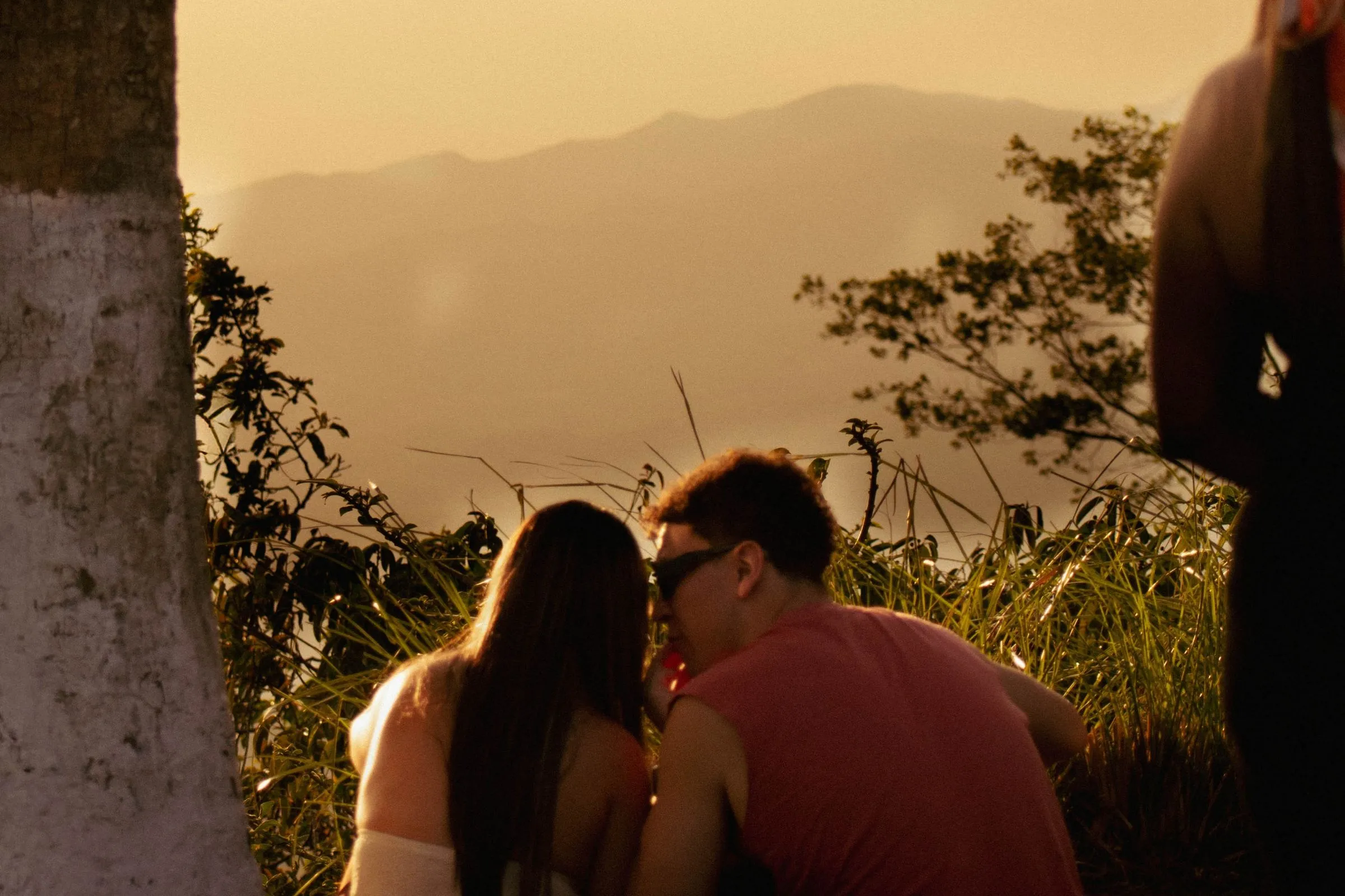 Una pareja sentada en el zacate mira el atardecer sobre los cerros de San Ramón