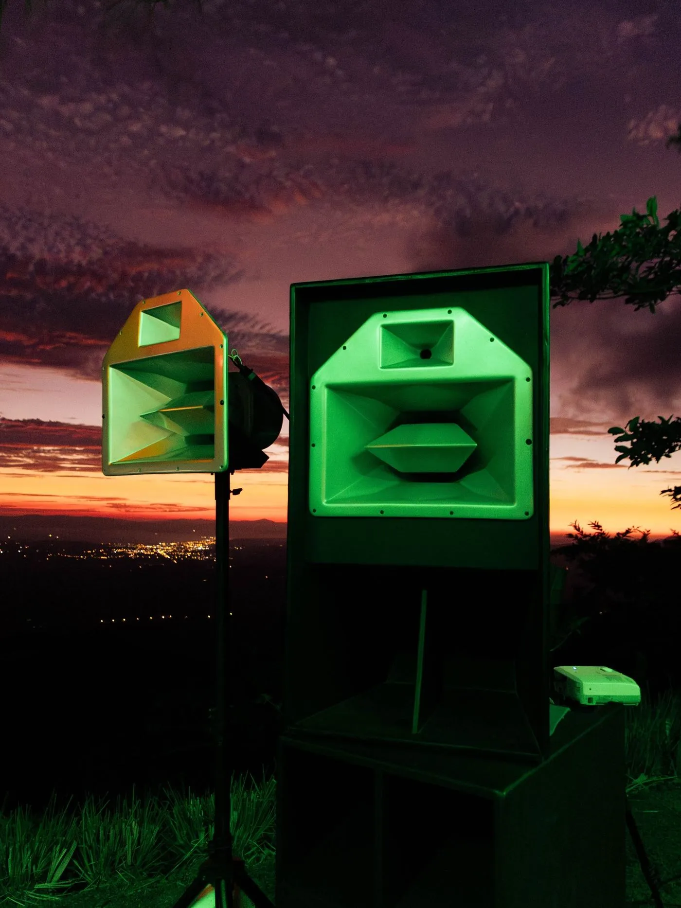 Dos parlantes iluminados de verde al anochecer, con el valle y las luces del pueblo atrás